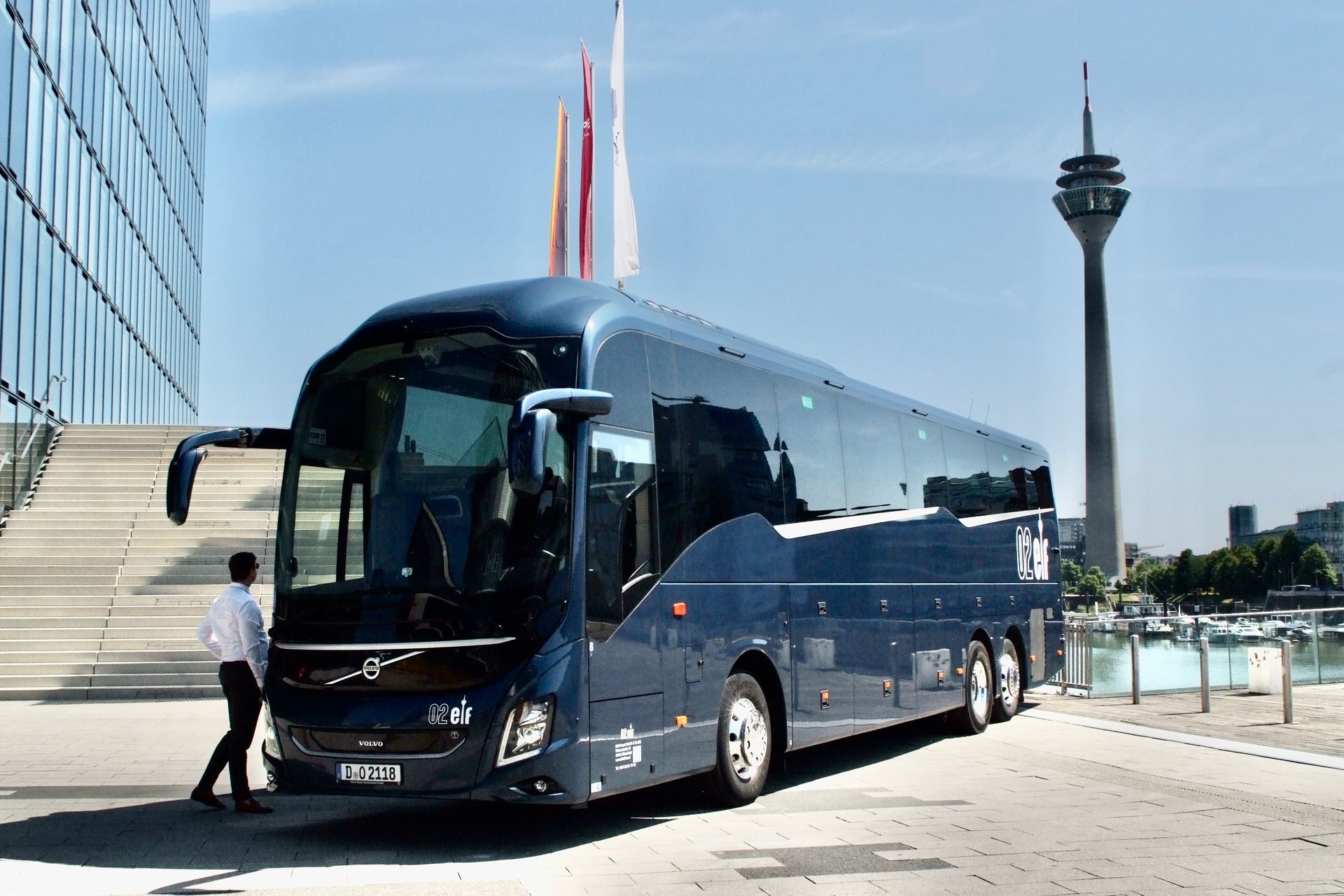Ein dunkelblauer Reisebus parkt neben einem modernen Gebäude, im Hintergrund ist ein Turm zu sehen.
