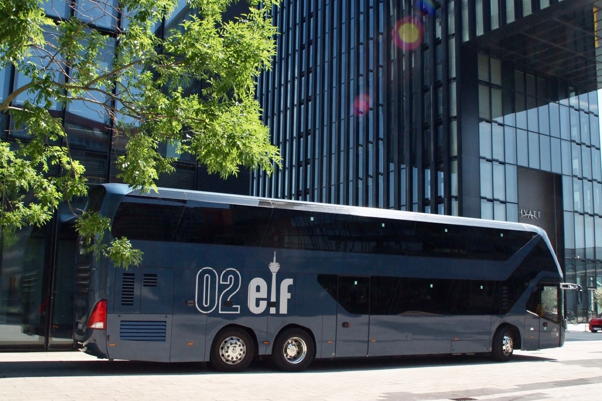 Ein dunkelgrauer elektrischer Reisebus parkt neben einem modernen Bürogebäude aus Glas, davor stehen Bäume.