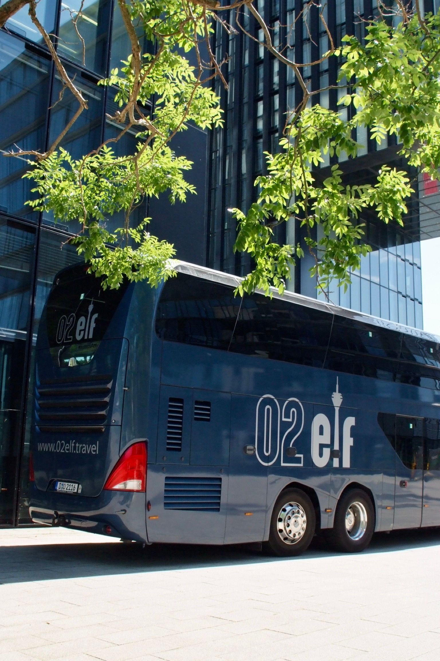 Ein blauer Doppeldecker-Reisebus parkt neben einem Glasgebäude unter Bäumen.