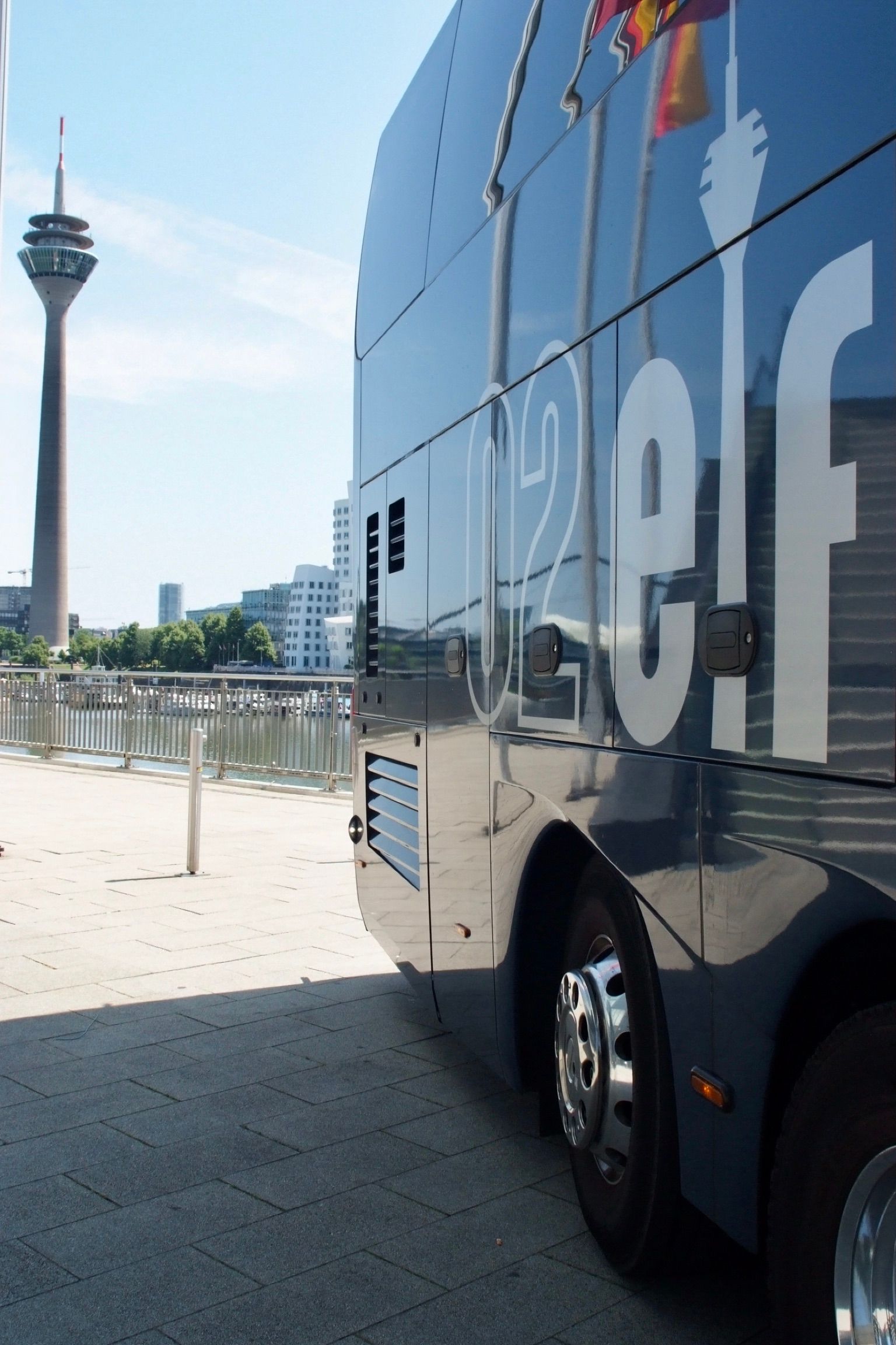 Ein reflektierender Reisebus parkt an einer Uferpromenade mit einem Turm im Hintergrund.