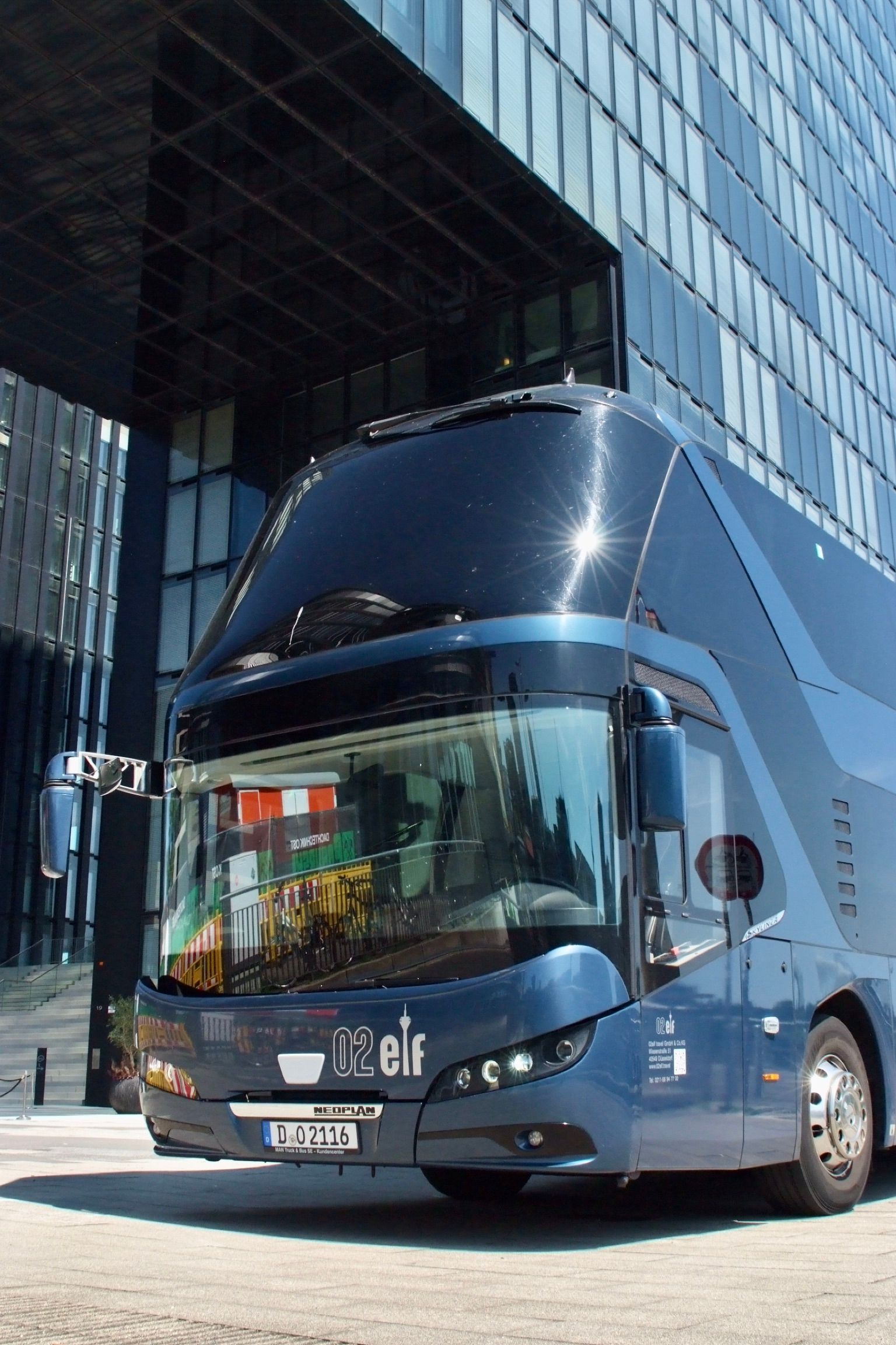 Ein blauer Doppeldeckerbus parkt neben einem gläsernen Bürogebäude in einer Stadtstraße.