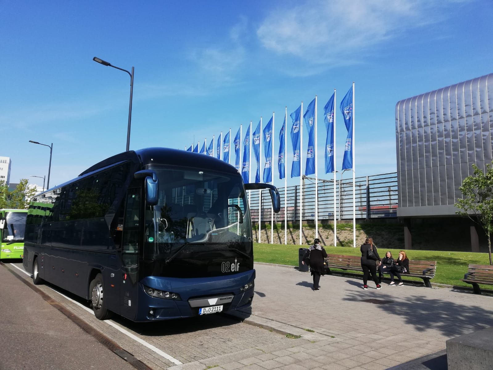 Ein dunkelblauer Reisebus parkt neben einem modernen Gebäude, in dem blaue Fahnen wehen und Fußgänger in der Nähe sind.
