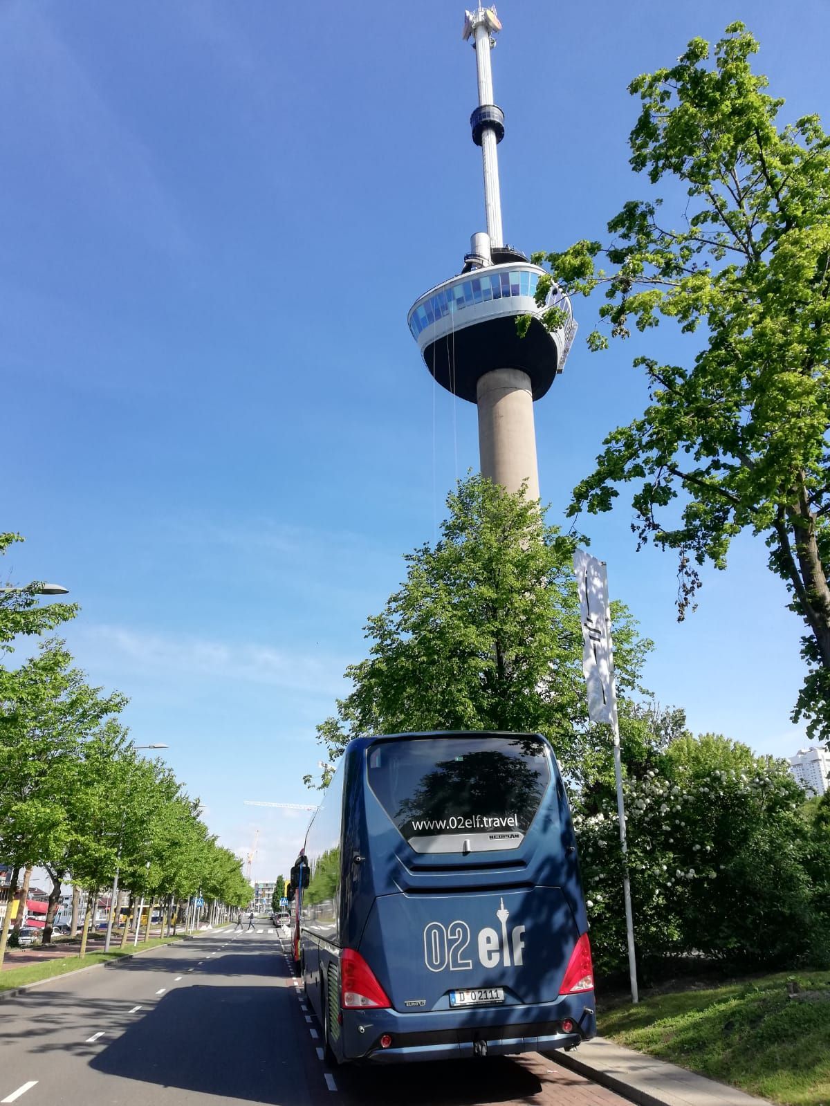 Ein blauer Reisebus auf einer von Bäumen gesäumten Straße, im Hintergrund ein hoher Turm unter klarem Himmel.