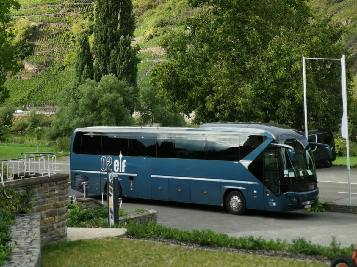 Ein blauer Reisebus auf einer Bergstraße neben einer Steinmauer und einem üppig grünen Hang.