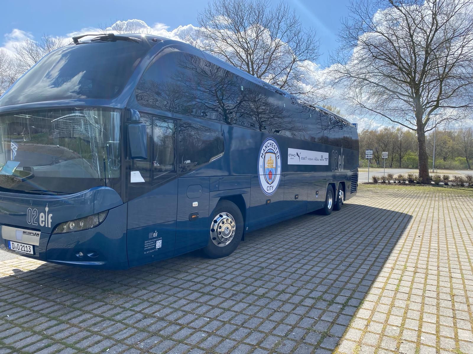 Ein dunkelblauer Reisebus parkt auf einem gepflasterten Parkplatz unter klarem Himmel; im Hintergrund sind Bäume zu sehen.