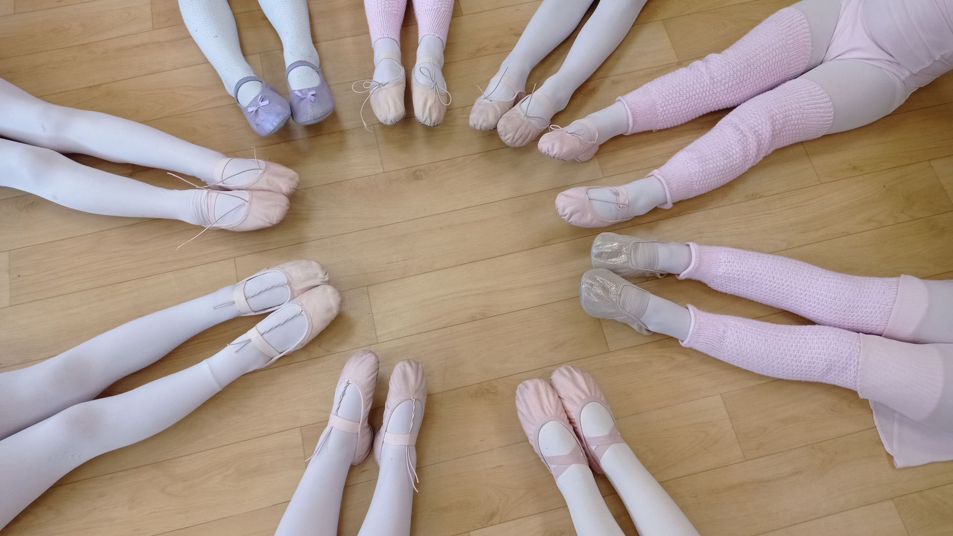 Piernas de bailarinas de ballet en círculo, con medias rosas y zapatillas de punta, sobre un piso de madera.