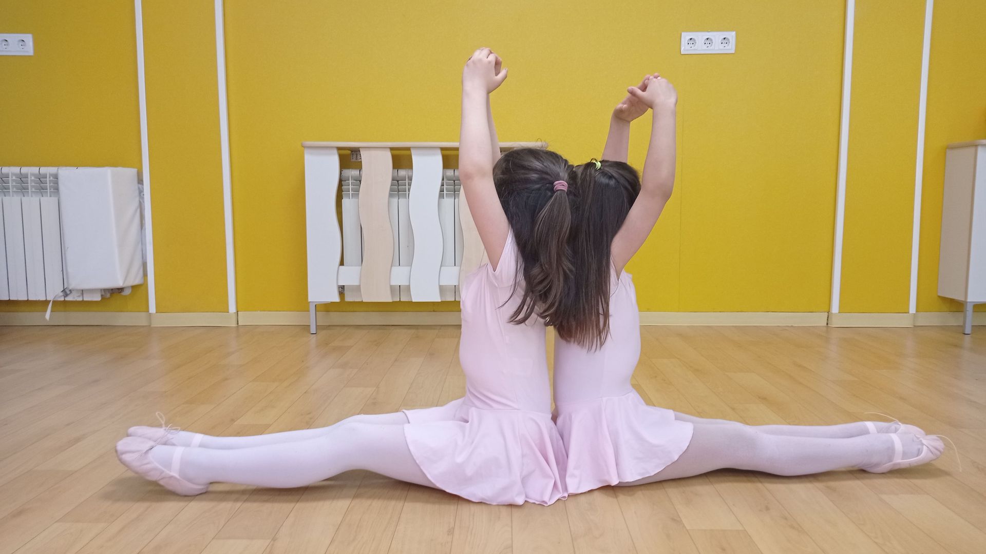 Dos bailarines vestidos de rosa haciendo el split con los brazos levantados, sobre un suelo de madera, en una habitación amarilla.