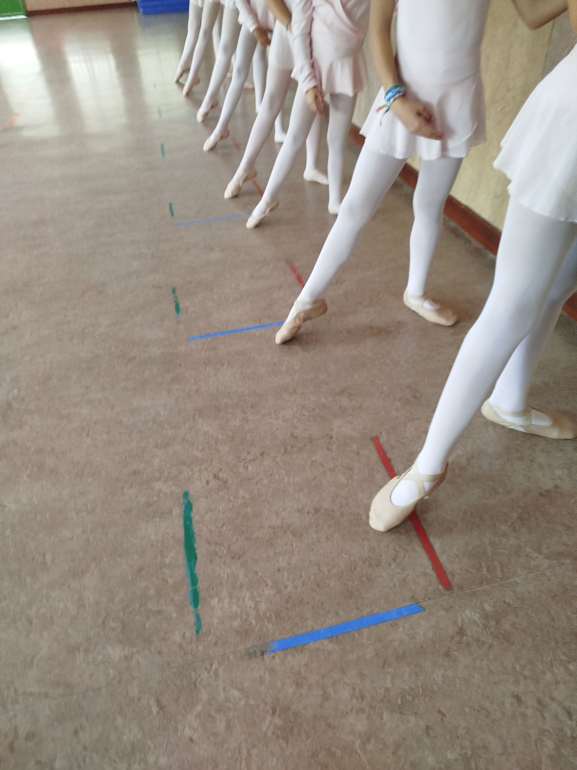 Bailarines de ballet vestidos de blanco, practicando poses con los pies sobre marcas en el suelo.