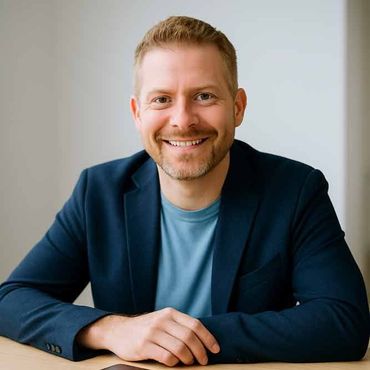 Lächelnder Mann in blauem Blazer und T-Shirt sitzt drinnen an einem Tisch.