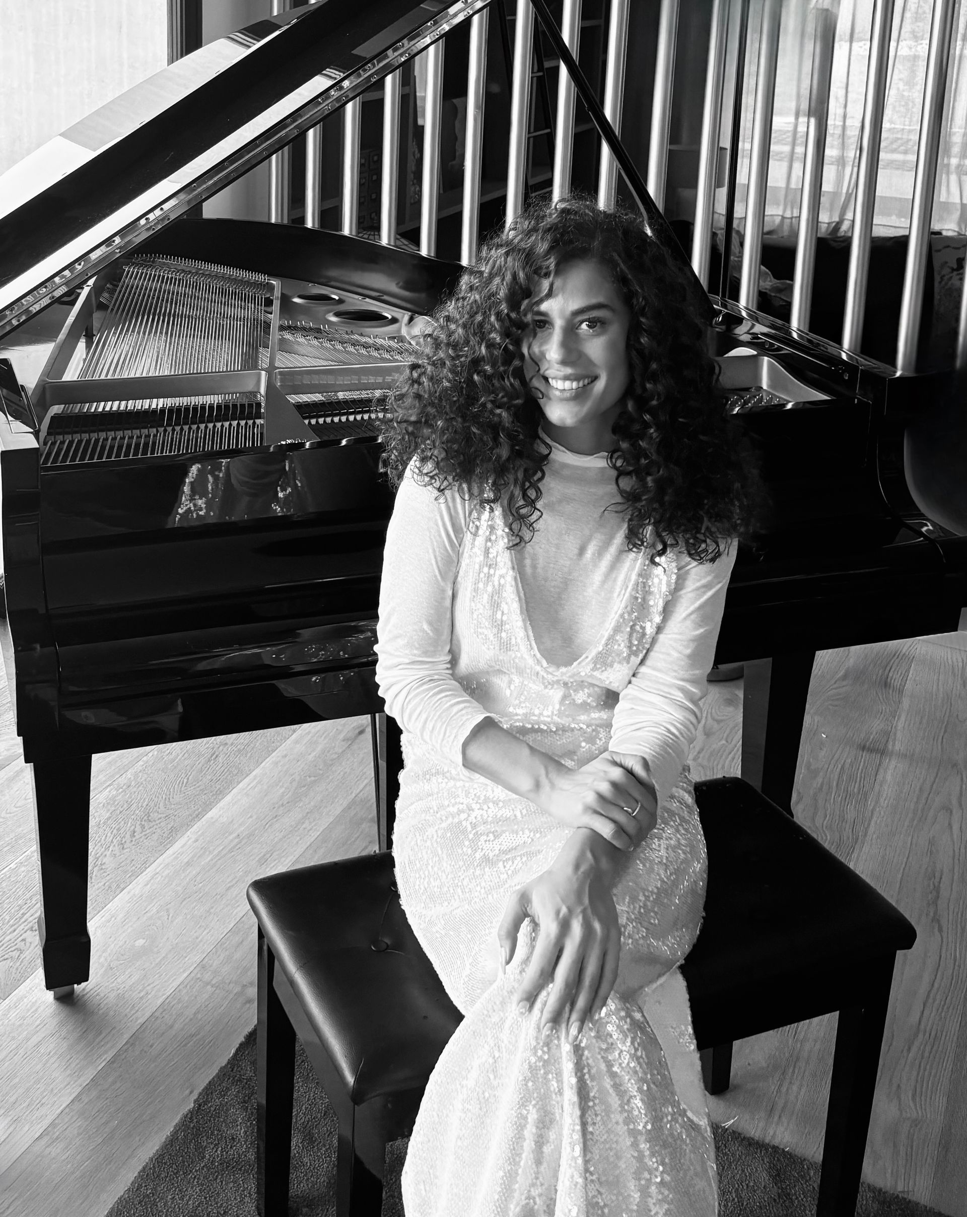 Una mujer vestida de blanco se sienta en el banco del piano, sonriendo, cerca de un piano de cola.