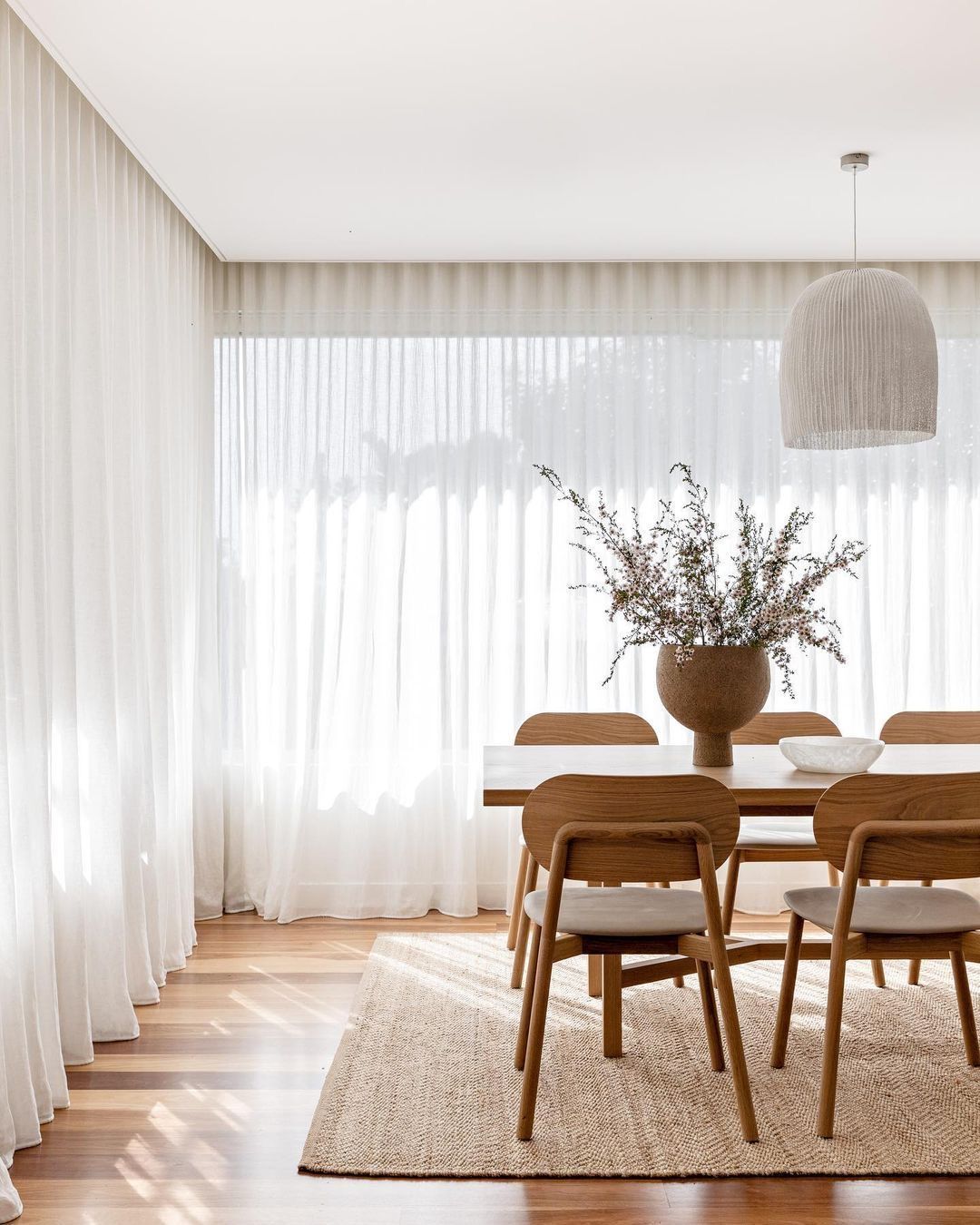 Comedor con mesa y sillas de madera, grandes cortinas blancas y alfombra beige.