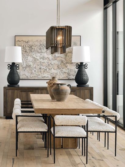 Comedor con mesa de madera, sillas, aparador, lámparas y obras de arte. Luz natural y colores neutros.