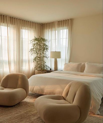 Acogedor dormitorio con cama, dos sillas curvas, cortinas beige y una planta alta bañada por la luz del sol.