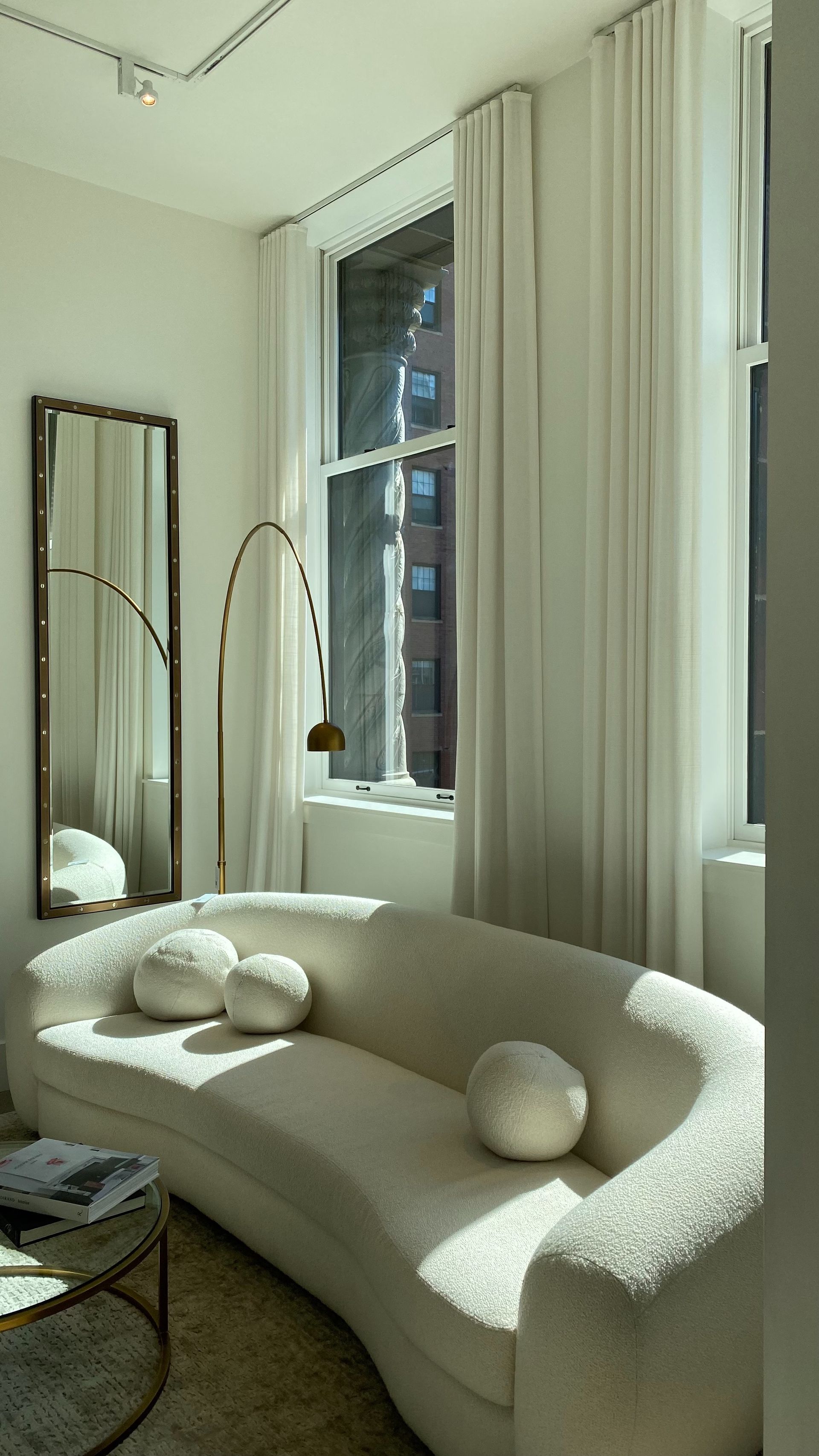 Acogedora y luminosa sala de estar con sofá curvo de color crema, detalles dorados y grandes ventanales con cortinas blancas.