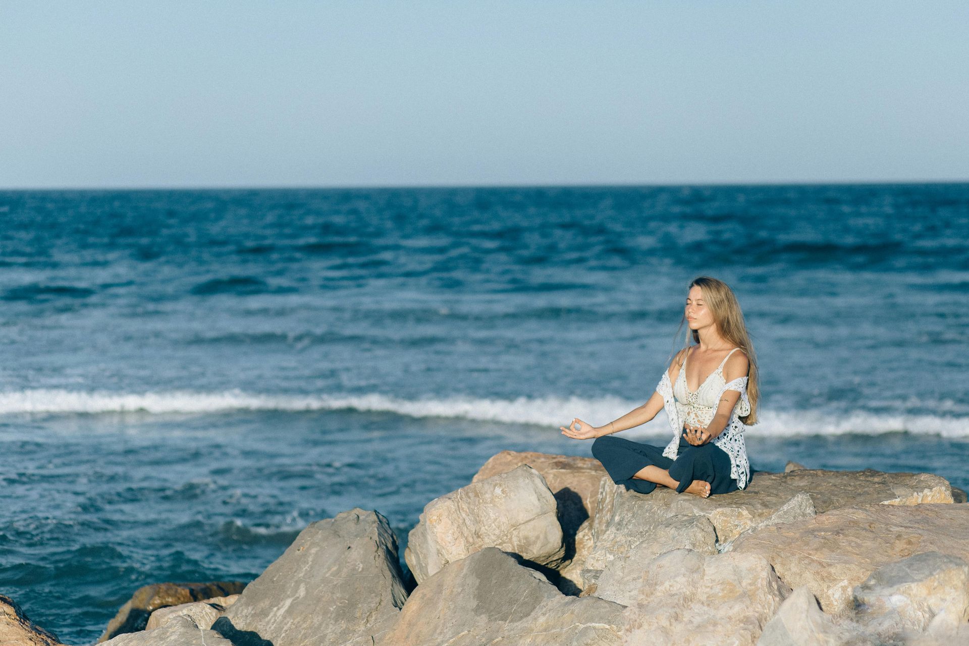 A woman is sitting on a rock near the ocean meditating.