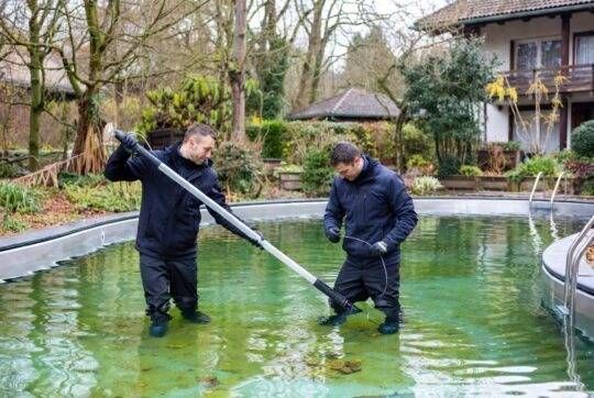 Zwei Personen reinigen im Freien einen grünen Pool mit langstieligen Werkzeugen.