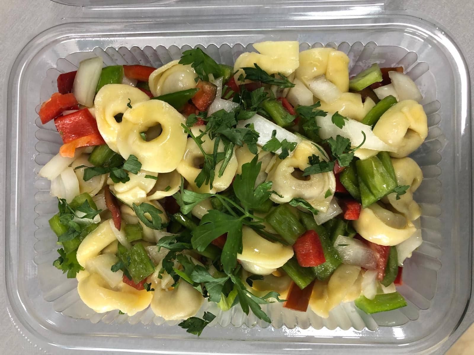 Ensalada de pasta tortellini con pimientos rojos y verdes, cebollas y perejil en un recipiente transparente.