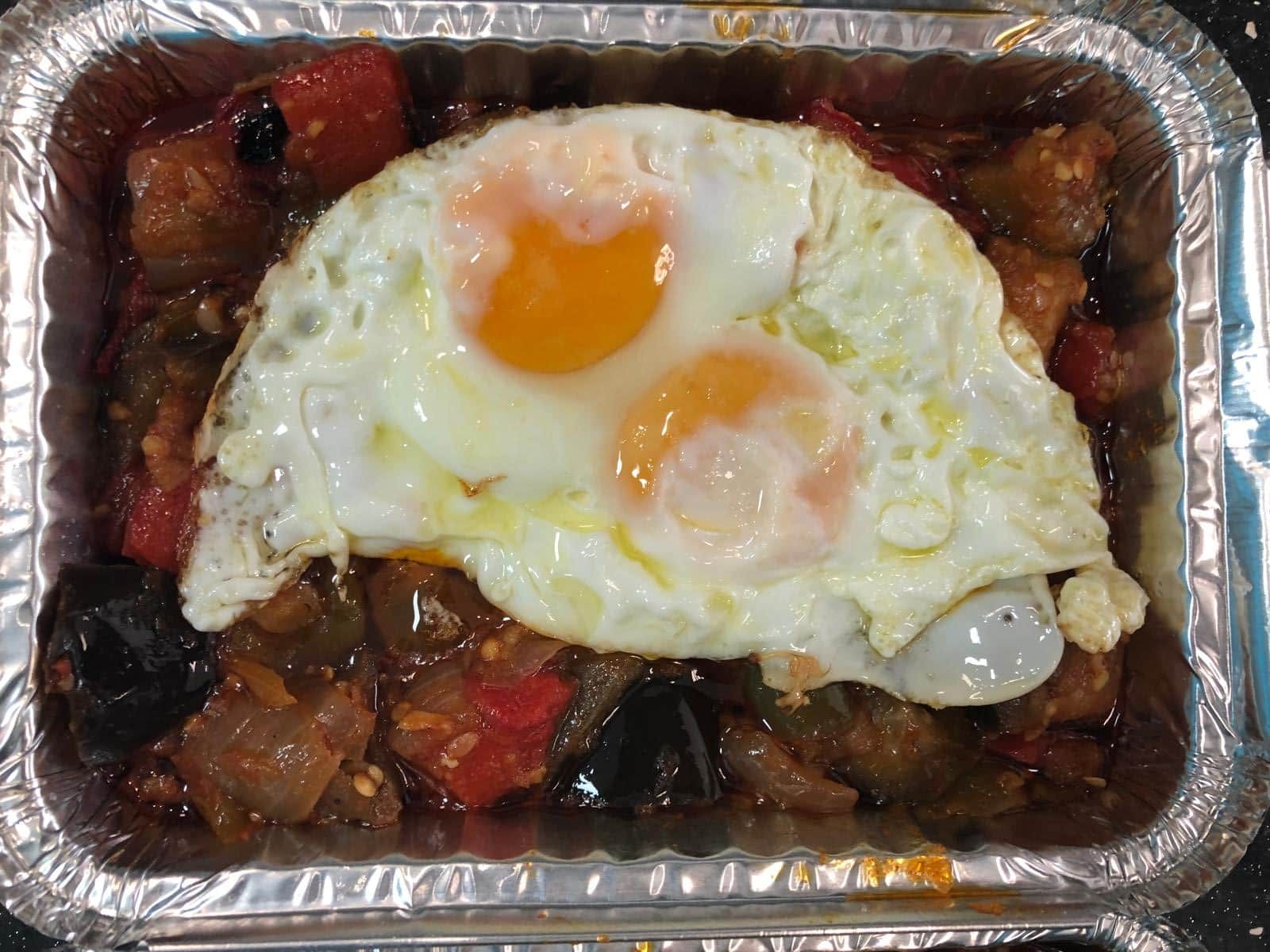 Plato de huevos y verduras en un recipiente de aluminio, huevos fritos encima de una mezcla de tomate y berenjena guisados.