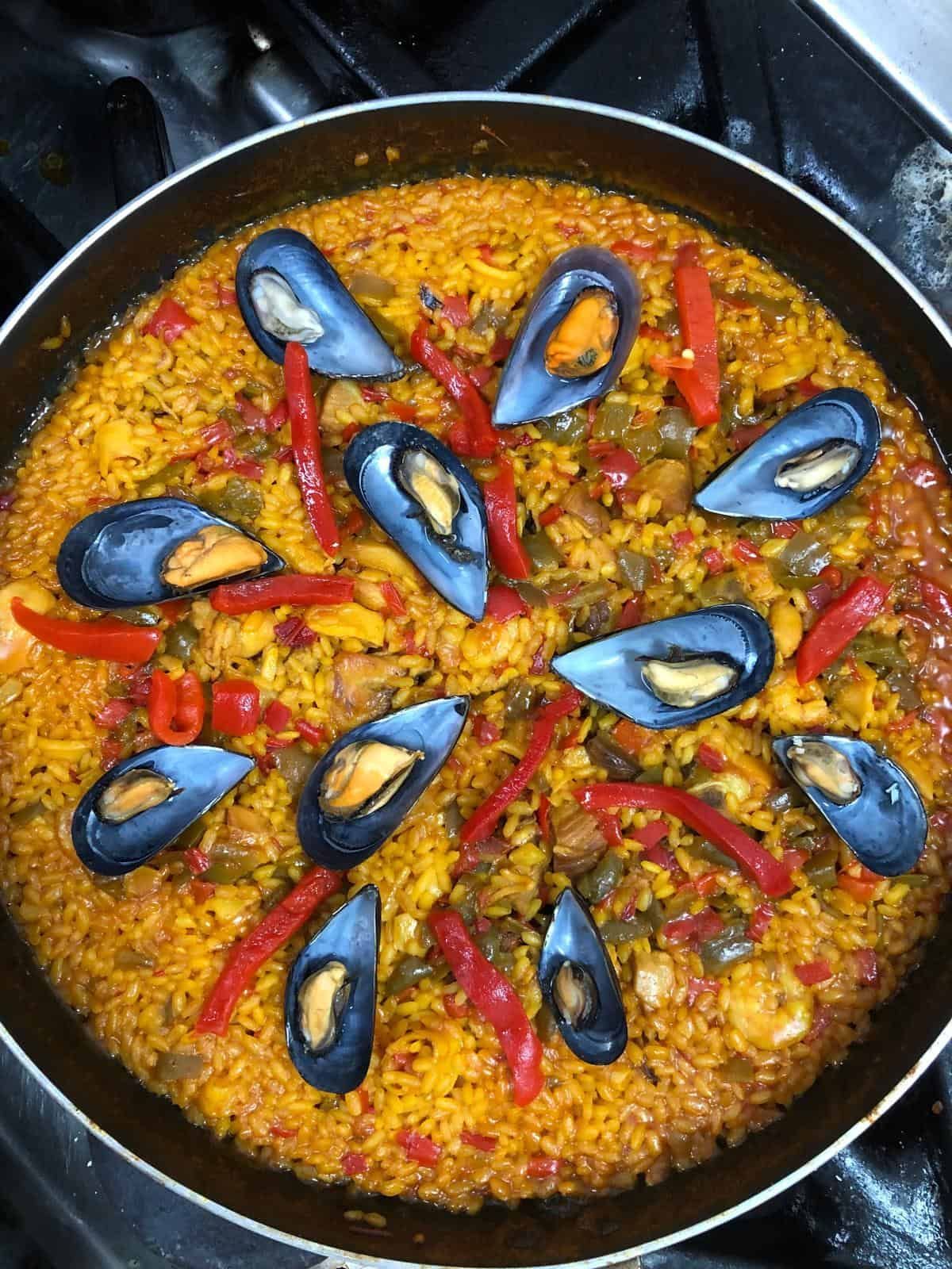 Paella con mejillones y pimientos rojos en sartén. Arroz amarillo con verduras anaranjadas y rojas.