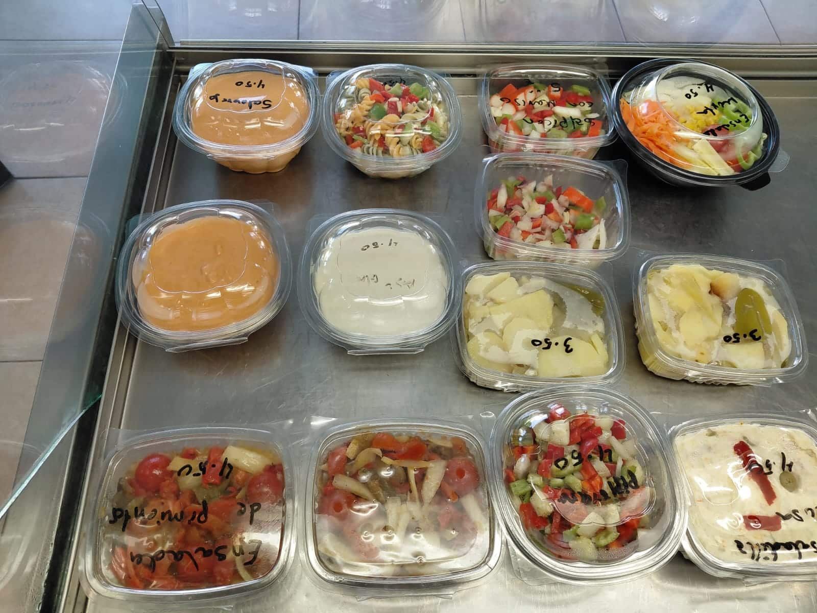 Varios recipientes de alimentos preenvasados ​​en una exhibición, que contienen ensaladas y salsas, en su mayoría transparentes y con etiquetas.