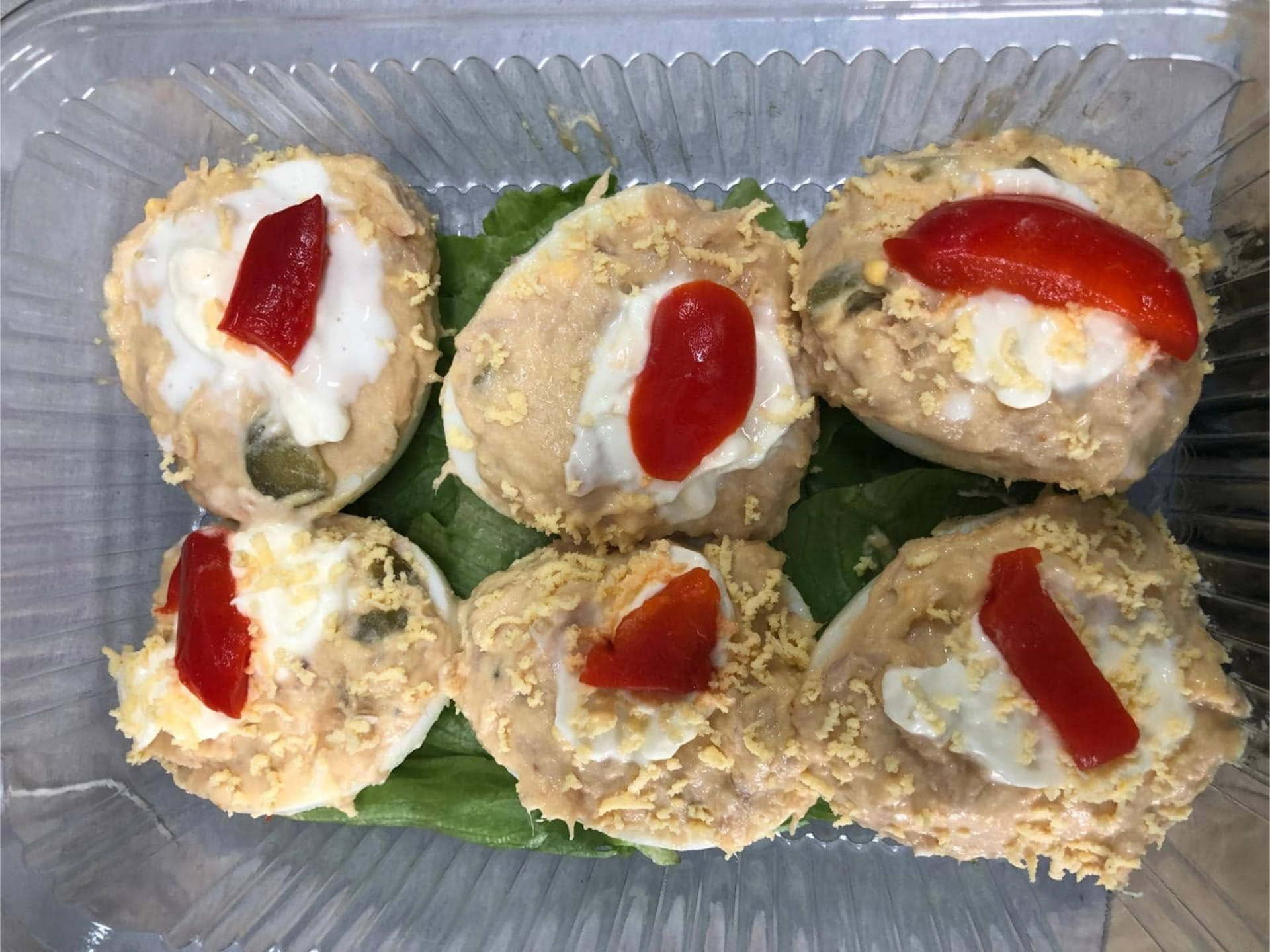 Huevos rellenos en un recipiente transparente, cubiertos con relleno blanco y pimiento rojo, sobre lechuga.