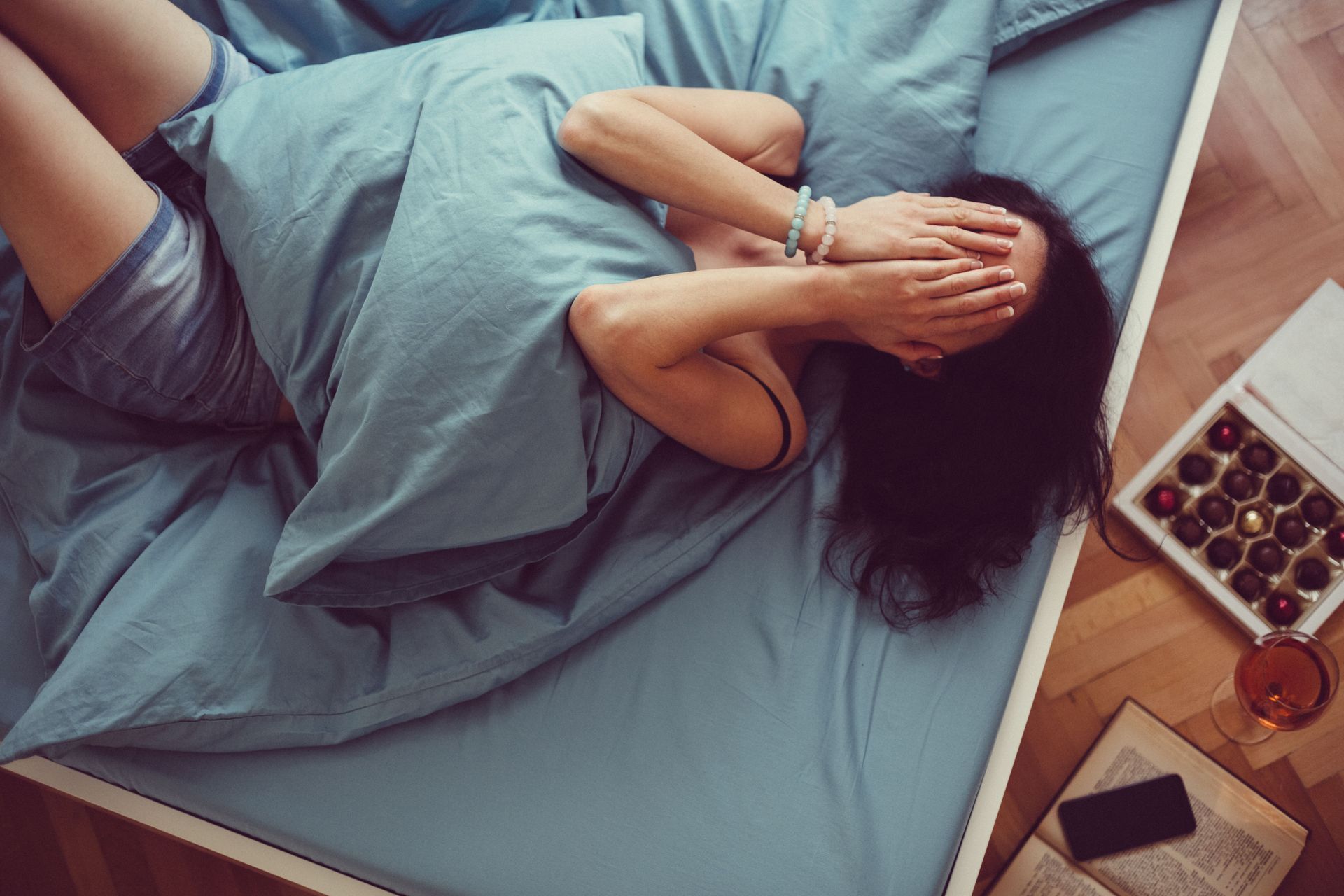 Mujer en una cama azul, cubriéndose el rostro con las manos. Chocolates, un libro y un teléfono en la mesita de noche.