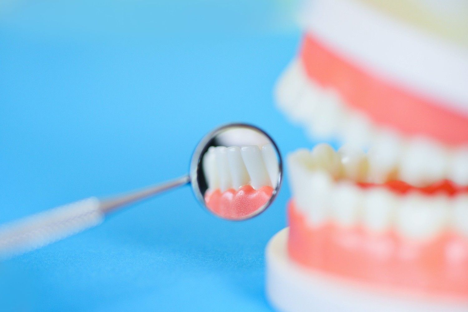 Espejo dental que refleja los dientes y las encías junto a un modelo dental sobre un fondo azul.