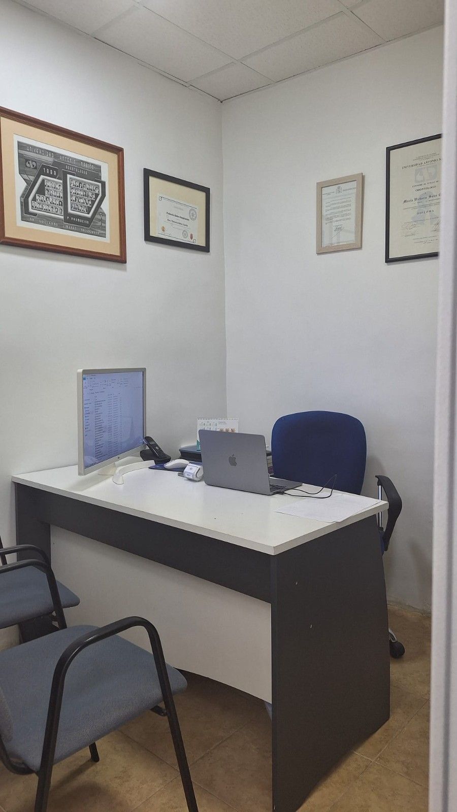 Pequeño rincón de oficina con un escritorio, una silla azul, un monitor de computadora y certificados.