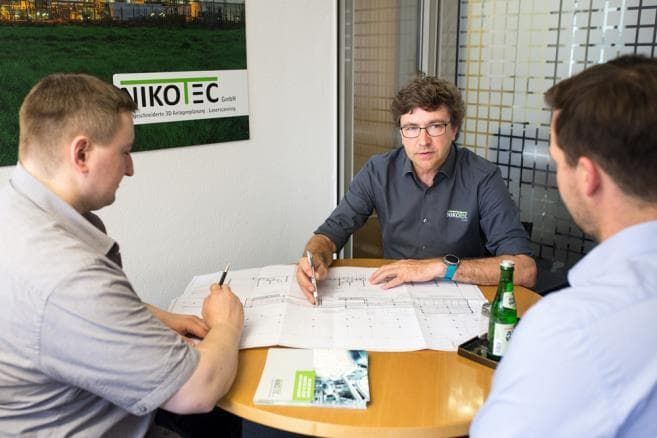 Drei Personen diskutieren an einem runden Tisch in einem Büro Dokumente; eine Flasche und Papiere sind im Blickfeld.