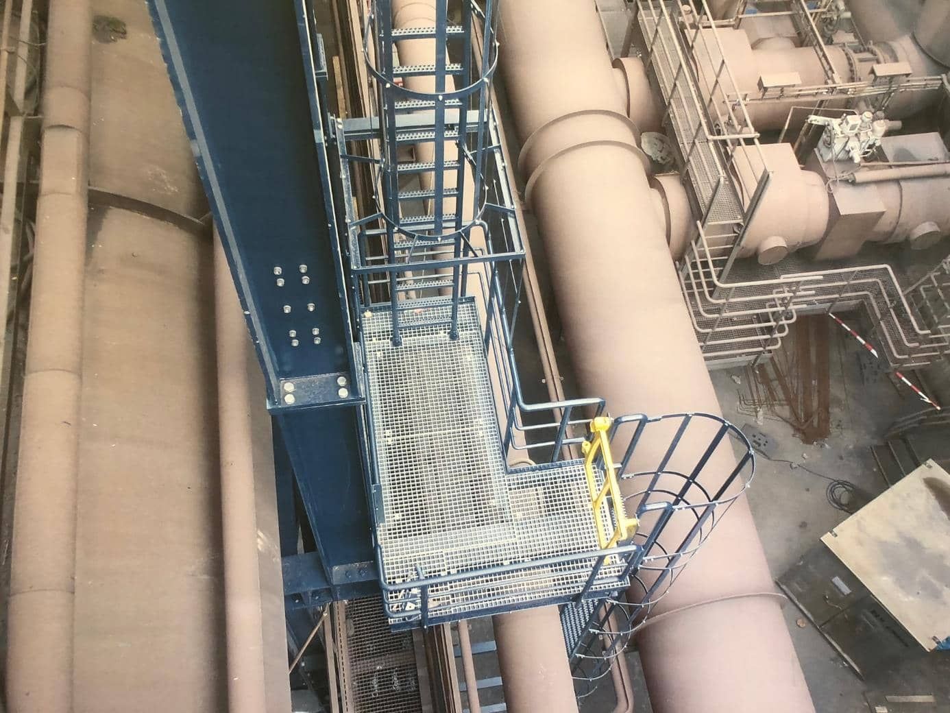 Industrieller Laufsteg neben großen hellbraunen Rohren und einer blauen Stahlkonstruktion, von oben betrachtet.