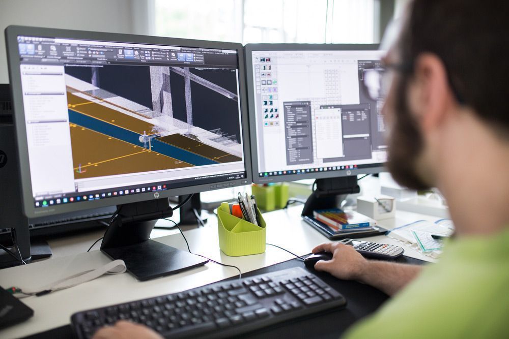 Person, die an einem Schreibtisch mit zwei Monitoren arbeitet, auf denen Designsoftware und ein 3D-Modell angezeigt werden.