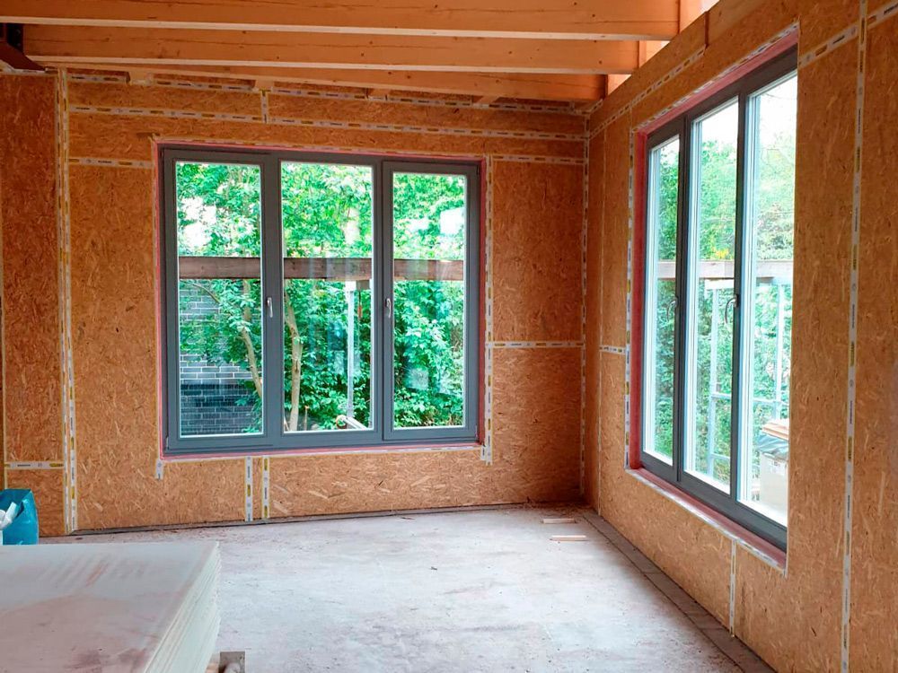 Unfertiger Raum mit freiliegendem Holzfachwerk, großen Fenstern und blankem Unterboden im natürlichen Licht