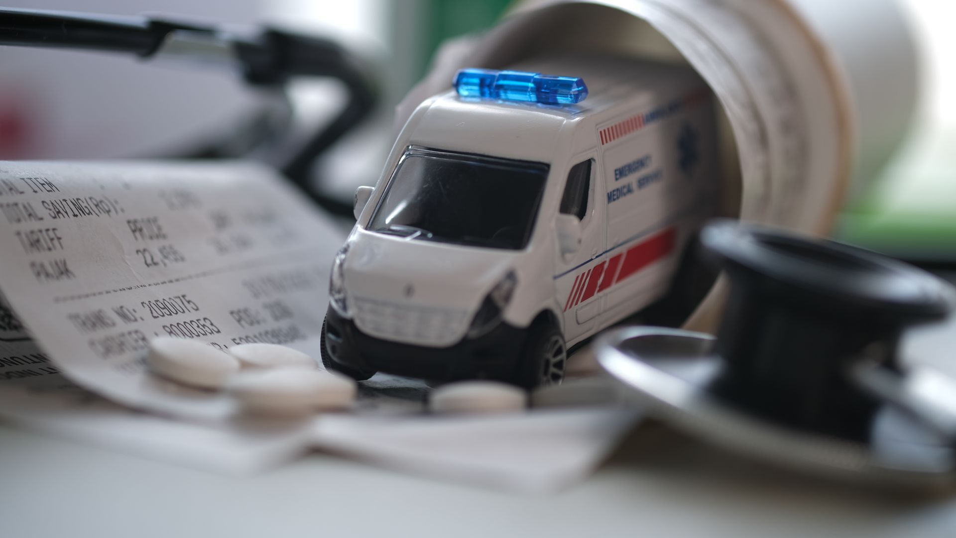 Une ambulance miniature posée sur un bureau à côté d'un stéthoscope, de pilules et d'un reçu bancaire.