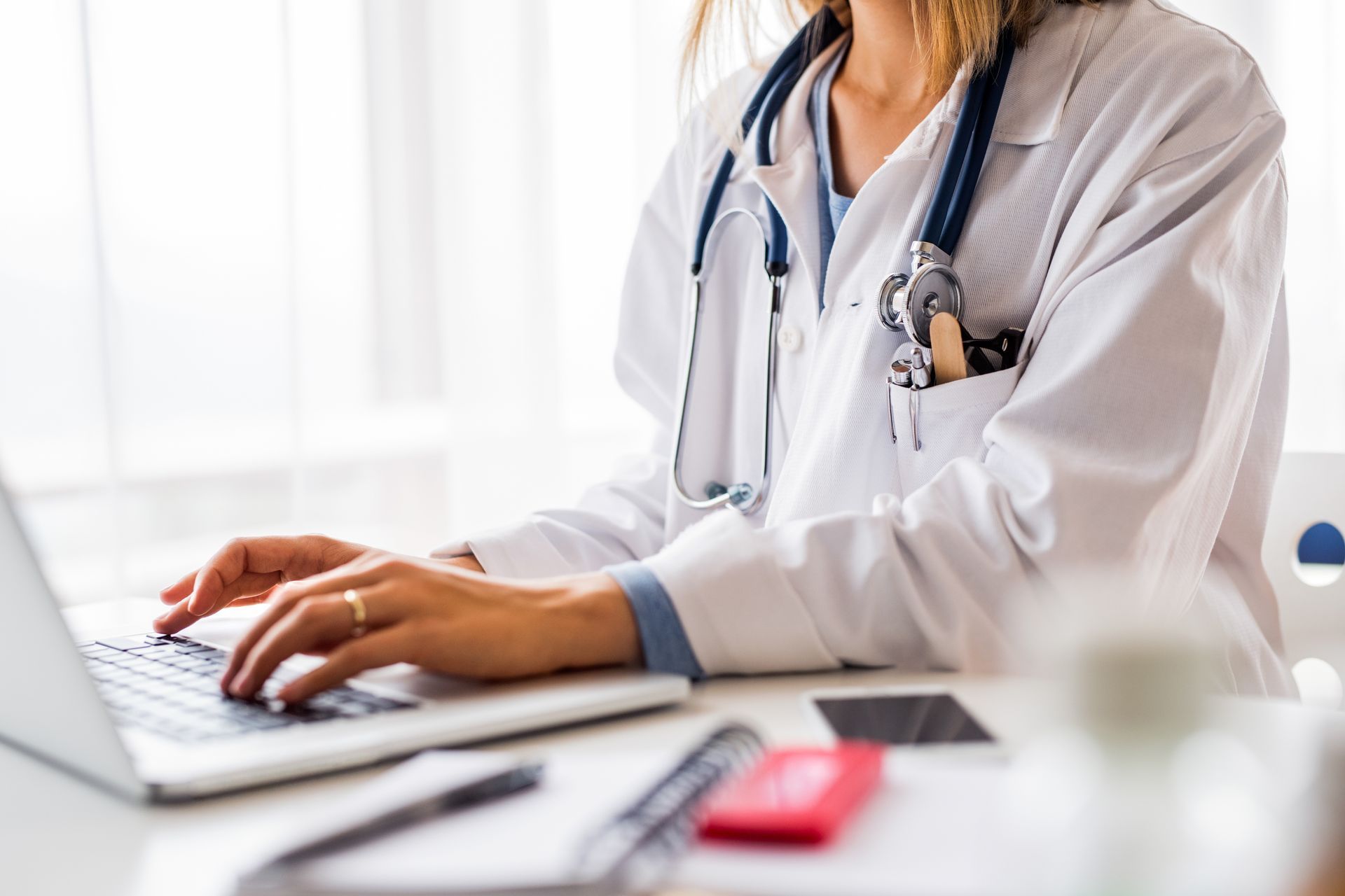 Un médecin tapant sur un ordinateur portable à son bureau, vêtu d'une blouse blanche et portant un stéthoscope.
