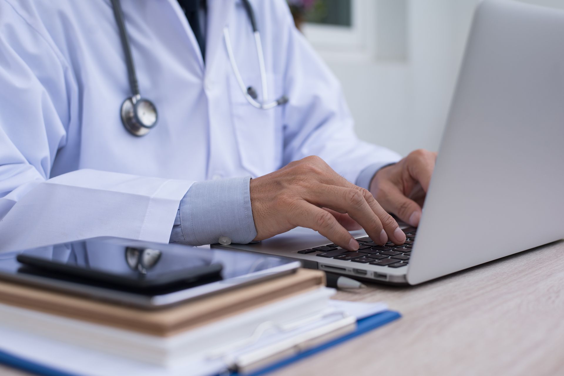 Un médecin tapant sur un ordinateur portable à son bureau, un stéthoscope autour du cou et un smartphone à proximité.