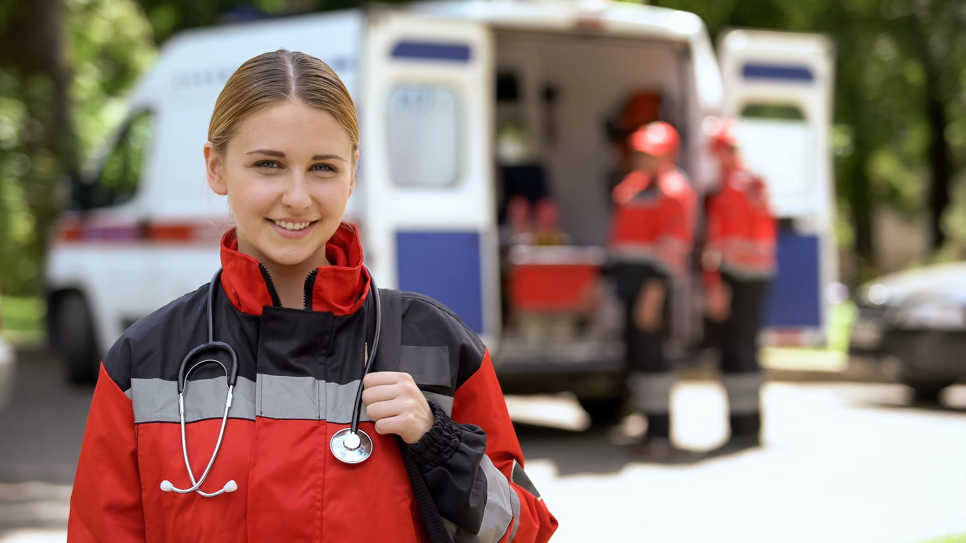 Une secouriste souriante en uniforme rouge et gris se tient devant une ambulance, stéthoscope autour du cou.