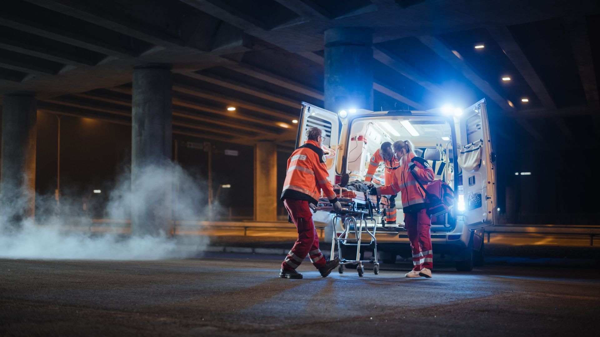 Des ambulanciers en uniforme orange chargent une civière dans une ambulance sous un pont autoroutier, de nuit.