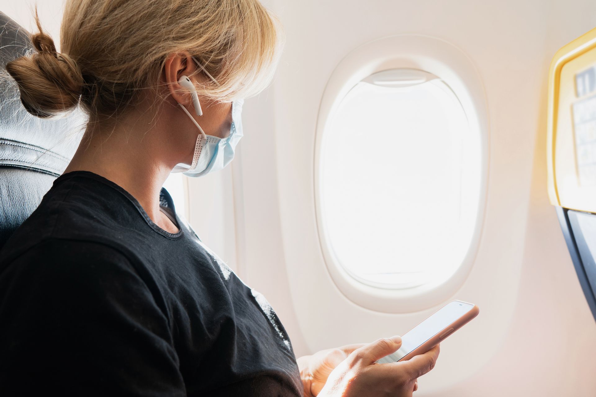 Une personne portant un masque regarde son smartphone près d'un hublot d'avion.