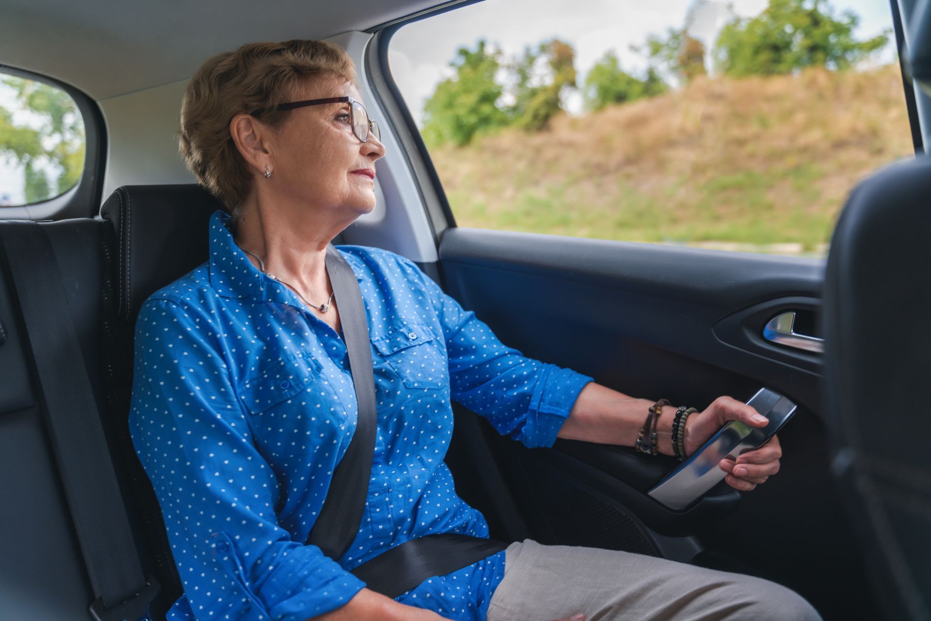 Une personne en chemise bleue, assise à l'arrière d'une voiture, se tient à la poignée et regarde le paysage.