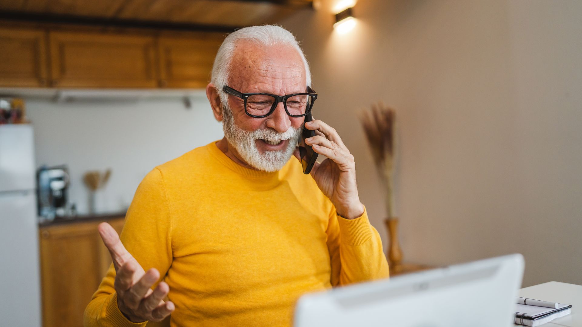 Un homme souriant, vêtu d'un pull jaune, parle au téléphone dans une cuisine.