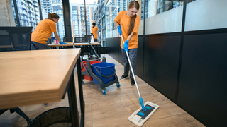 Una enfermera consuela a una anciana, sujetándole la mano y el brazo. Ambas están sentadas en el interior, vestidas con ropa profesional.