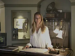 Una mujer con camisa blanca sonríe, de pie detrás de un escritorio con lámpara, libros y objetos antiguos en una habitación con poca luz.