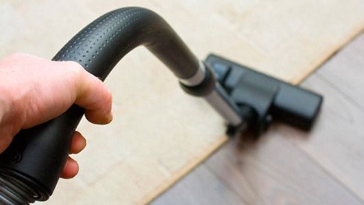 Mano sosteniendo una manguera de aspiradora, limpiando una zona alfombrada junto a un suelo de madera.