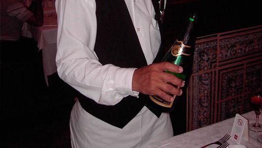Camarero con camisa blanca y chaleco negro sosteniendo una botella de champán verde en una mesa.