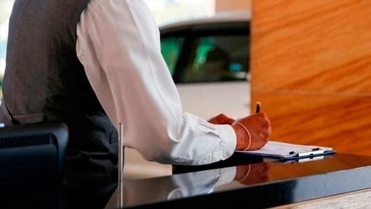 Persona sentada en un escritorio escribiendo en una tabla con portapapeles, posiblemente en el registro de entrada de un hotel.