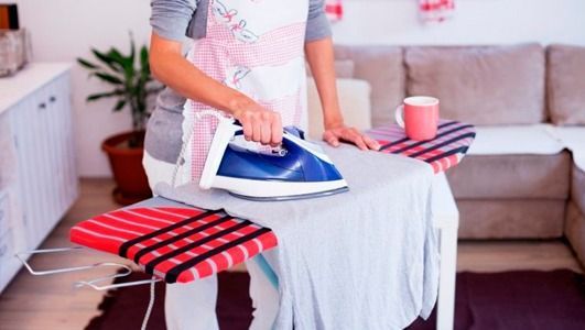 Una mujer plancha una camisa gris claro sobre una tabla de planchar en una sala de estar; cerca hay un asiento a rayas rojas.