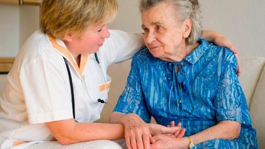 Una persona mayor, acompañada de consuelo por un cuidador, se encuentra sentada en el interior de una vivienda.