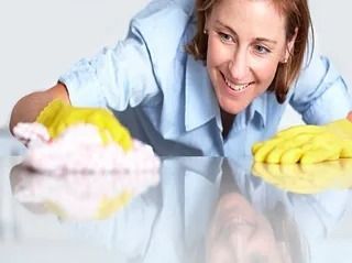 Mujer sonriendo mientras limpia una superficie reflectante con una mano enguantada de amarillo y un paño rosa.