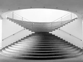 Escalera curva que asciende hasta una abertura circular, con dos tramos que se cruzan en el aire.