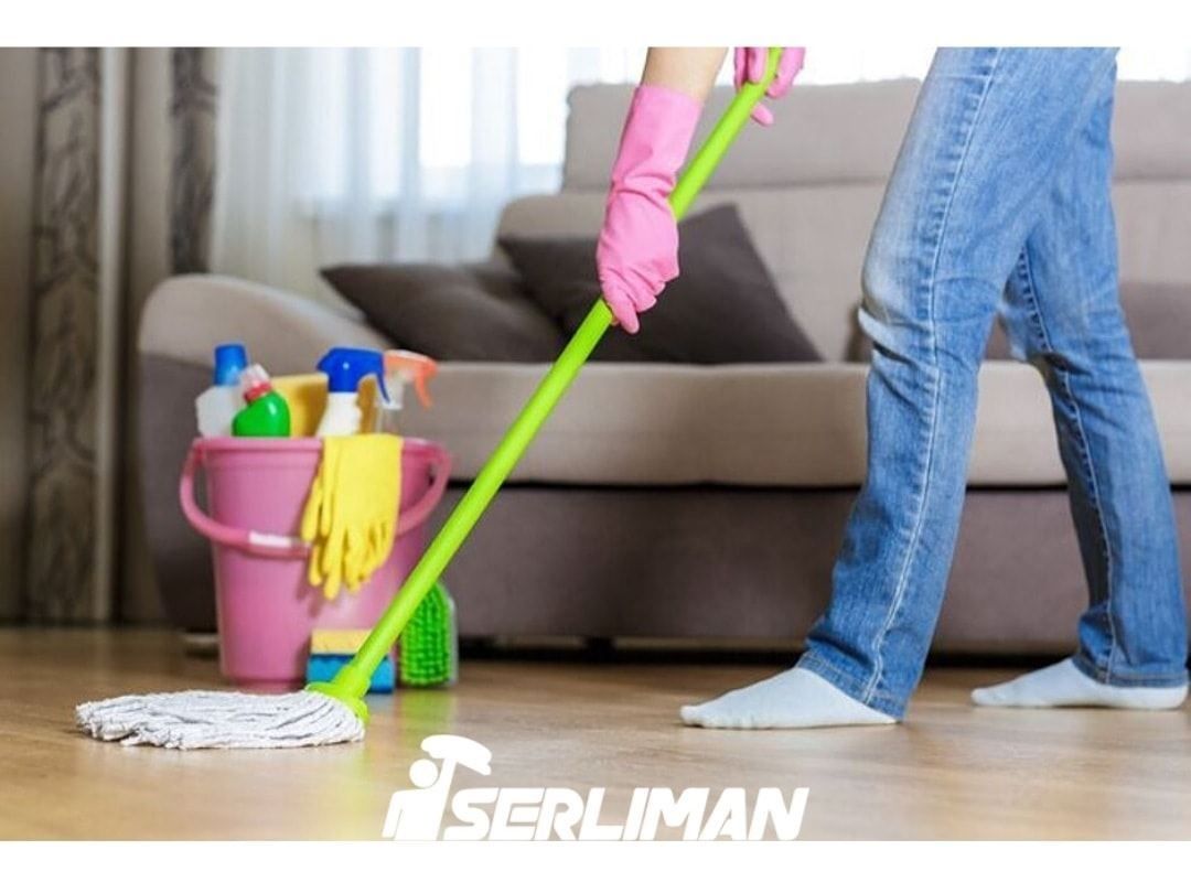 Persona fregando un suelo de madera; productos de limpieza en un cubo rosa junto a un sofá beige.